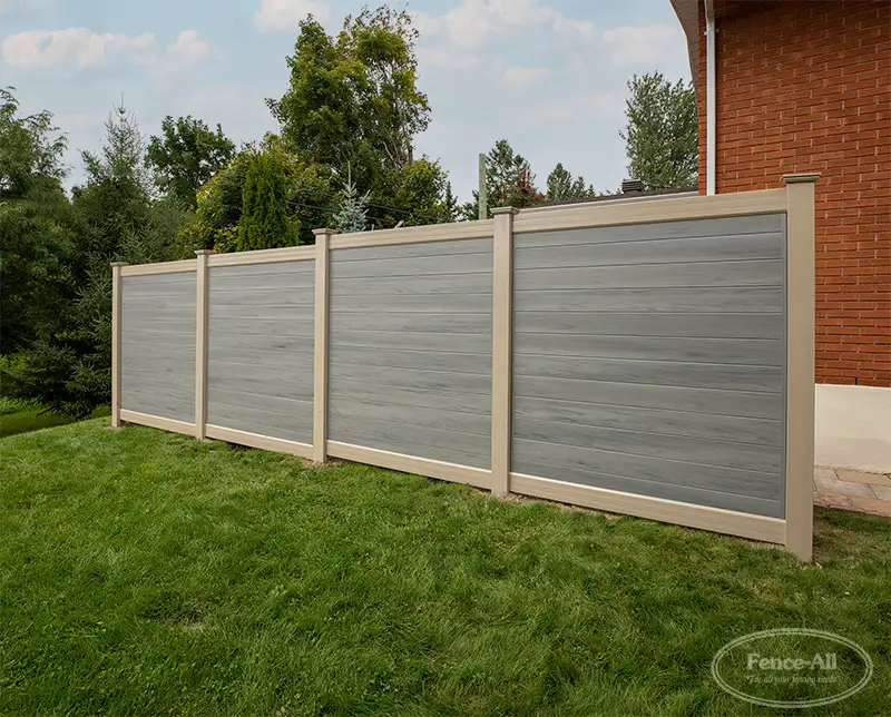 A two-tone privacy fence with horizontal grey wood-grain panels and tan posts stands on a green grass lawn. The fence is positioned next to a red brick house, with trees and a lightly cloudy sky in the background.