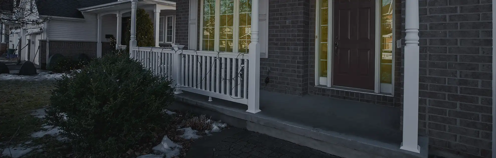 A residential front porch with dark brick siding featuring a white vinyl railing and matching support pillars. String lights are woven through the balusters, with a large green shrub and patches of snow in the foreground.
